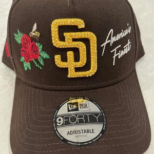 May include: Brown baseball cap with the text "American's Finest" and a gold-colored "SD" logo. The cap features a floral and bee design on the side. The hat is adjustable and has a "9FORTY" label.