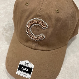 May include: A brown baseball cap with a bedazzled "C" in the center. The letter is outlined with clear rhinestones and filled with copper-colored rhinestones. The cap has a curved brim and an adjustable strap. A tag on the brim reads "OSFA ADJUSTABLE".