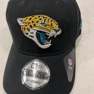 May include: Black adjustable baseball cap featuring a jeweled jaguar logo in gold, teal, and silver. The cap has a curved brim and a New Era 9TWENTY adjustable sticker. A small NFL Team tag is attached.