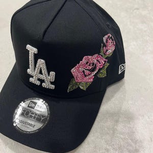 May include: Black baseball cap with the letters "LA" in silver rhinestones on the front. A pink rhinestone rose design is on the side. The cap has a curved brim and an adjustable strap.
