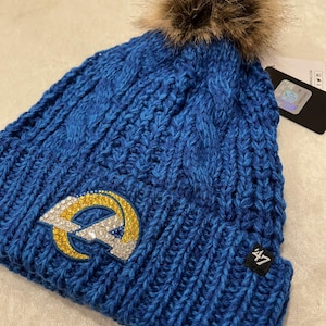 May include: Blue knitted beanie with a faux fur pom pom on top. The beanie has a large, sparkly, blue, gold, and white logo on the front. The logo is the Los Angeles Rams logo. The beanie has a 47 brand tag on the side.