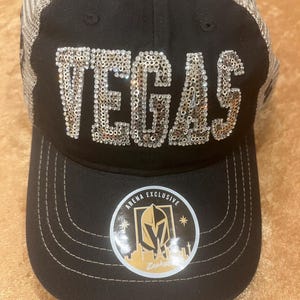 May include: A black baseball cap with a mesh back and the word VEGAS spelled out in silver sequins across the front. A circular sticker on the brim features a gold knight helmet logo, the text ARENA EXCLUSIVE, and a city skyline silhouette.