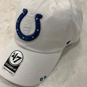 May include: White adjustable baseball cap with a blue horseshoe logo embellished with rhinestones. The cap features the text "47 CLEAN UP ADJUSTABLE" on the brim and small blue flower accents.