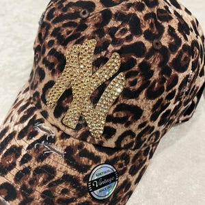 May include: A brown and black leopard print baseball cap with a large gold rhinestone NY logo. The cap has a distressed look with holes and frayed edges. The cap has a small black and silver label that says "KBETHOS Vintage Since 2001".