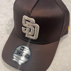 May include: A brown baseball cap with a curved brim. The cap features a sparkling "SD" logo on the front. The adjustable hat has a "9FORTY" sticker on the brim.