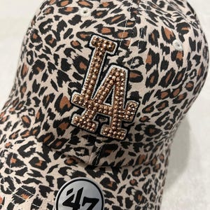 May include: A baseball cap with a leopard print pattern in tan, brown, and black. The front features the letters "LA" in gold with rhinestone embellishments. A tag with the number "47" is visible.