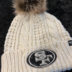May include: White knitted beanie with a faux fur pom pom and a black and white rhinestone San Francisco 49ers logo.