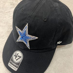 May include: A black baseball cap featuring a star emblem on the front panel, embellished with blue and clear rhinestones. The cap includes a 47 brand logo on the side and a circular 47 Clean Up Adjustable sticker on the curved brim.