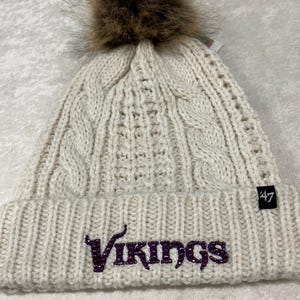 May include: White knitted beanie with a brown faux fur pom pom and a purple "Vikings" logo with rhinestones. The beanie has a 47 brand label.