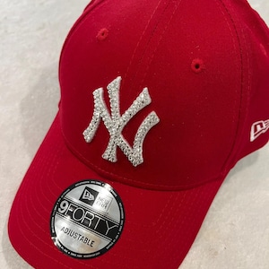 May include: A red adjustable baseball cap with a white, rhinestone-studded "NY" logo. The cap features a curved brim and a small New Era logo on the side. The adjustable strap is visible, and a "9FORTY" sticker is on the brim.