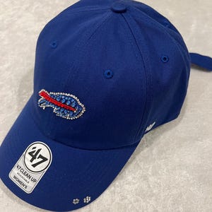 May include: A royal blue baseball cap featuring a Buffalo Bills logo embellished with red, blue, and clear rhinestones on the front panel. The brim displays a white sticker with the text 47 CLEAN UP ADJUSTABLE and two small rhinestone flower accents.