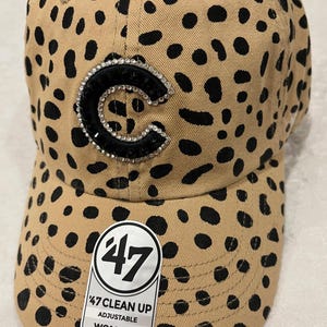 May include: Tan baseball cap with black spots and a rhinestone-studded "C" logo. The cap features a curved brim and an adjustable strap. The tag reads "47 CLEAN UP ADJUSTABLE WOMEN'S".