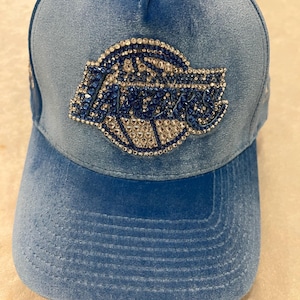 May include: A light blue velvet baseball cap featuring the word LAKERS in a stylized font. The logo is embellished with blue and clear rhinestones that create a sparkling effect across the front panels of the hat, which has a curved brim.