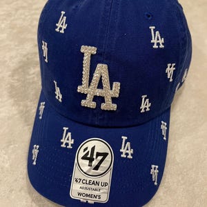 May include: A royal blue baseball cap with the letters "LA" in white embroidered across the front. The cap has a curved brim and features the "LA" logo in rhinestones. The tag reads "47 Clean Up Adjustable Women's".