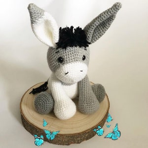 May include: A crocheted gray and white donkey with black yarn for the mane and tail. The donkey is sitting on a wooden slice with blue butterflies around it.