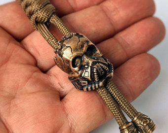 May include: A brown and green paracord bracelet with a bronze skull charm. The skull has a detailed design with a gas mask.