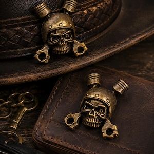 Puede incluir: Dos emblemas metálicos idénticos en tono latón que muestran una calavera con casco, flanqueada por dos pistones de motor en la parte superior y dos bielas en la parte inferior. Los emblemas se muestran sobre un sombrero y una cartera de cuero marrón.