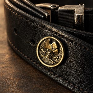May include: A black leather belt featuring a circular brass-toned emblem with a raised skull and crossbones design. The belt includes a metallic buckle mechanism and visible stitching along the edges of the strap.