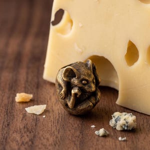 Pode incluir: Uma pequena conta metálica de cor bronze em forma de rato enrolado, com textura de pelo detalhada e orelhas pontiagudas. A figura do rato está posicionada em frente a um grande pedaço de queijo amarelo com buracos visíveis.