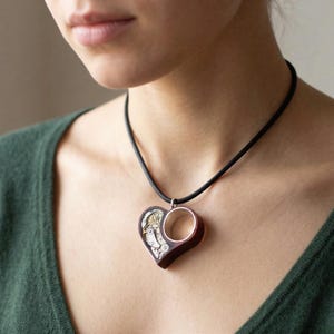 Könnte beinhalten: Halskette mit herzförmigem Anhänger aus dunklem Holz und Silber. Der Anhänger hat einen kreisförmigen Silberakzent und ein detailliertes mechanisches Uhrwerk-Design. Die Halskette hat ein schwarzes Band.