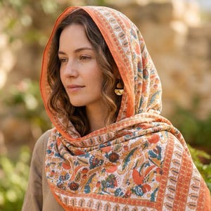 May include: A patterned scarf with an orange border, worn over the head and shoulders. The scarf features a detailed floral design in blue, orange, and white. The background shows a natural outdoor setting with plants and a stone wall.