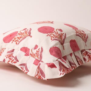 May include: A decorative pillow with a white background and a repeating pattern of pink floral designs. The pillow has a ruffled edge and is designed for home decor.