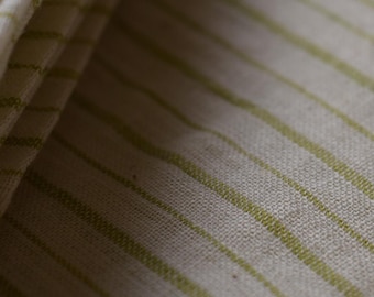 May include: Close-up of folded fabric with a cream-colored base and thin, parallel green stripes. The fabric is textured and appears to be linen or a similar natural fiber. The stripes run diagonally across the material.