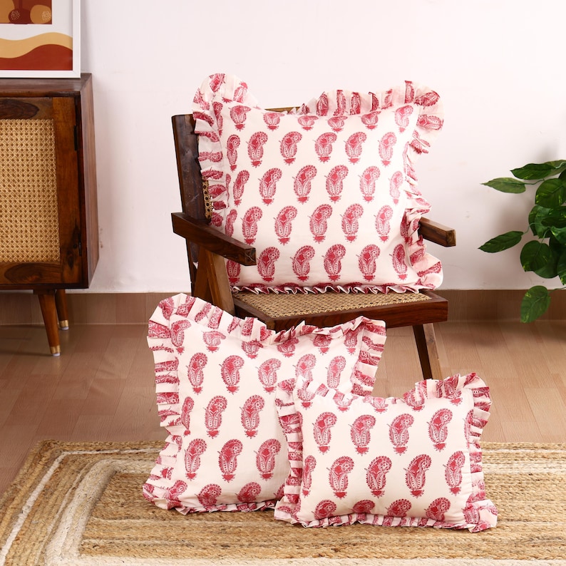 May include: Three square cushion covers with a white background and a red paisley pattern. The cushion covers have a white frilled trim around the edges. The cushion covers are sitting on a woven rug.