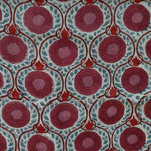 Op de afbeelding: Een stof met een herhalend bloemenpatroon. Het ontwerp toont grote, cirkelvormige bloemen in tinten bordeauxrood en roze, tegen een crèmekleurige achtergrond. De bloemen worden omlijst door een donkerbruine, gebogen omtrek met blauwgroene bladeren.