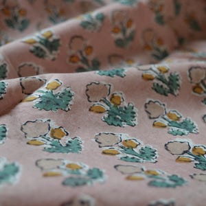 May include: Close-up of a pink fabric with a repeating floral pattern. The design features light beige flower shapes with yellow centers and teal green leaves. The fabric is slightly wrinkled, suggesting it is soft.