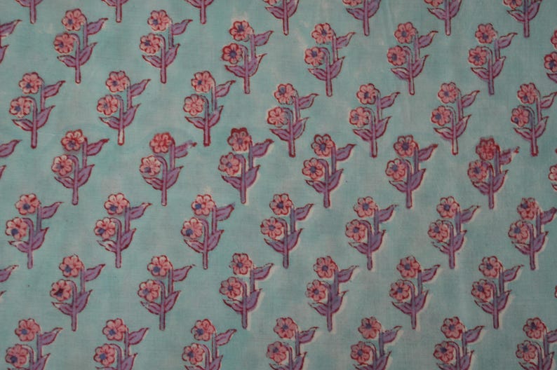 May include: A repeating pattern of pink flowers with purple stems and leaves on a light blue background. The floral design is evenly spaced across the fabric, creating a visually appealing textile pattern. The flowers have multiple petals.