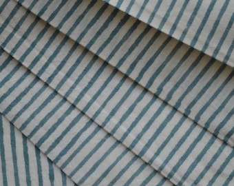 May include: Close-up of folded fabric with a repeating pattern of teal blue stripes on an off-white background. The stripes are uneven and hand-drawn in appearance. The fabric is folded in a diagonal pattern, creating a layered effect.