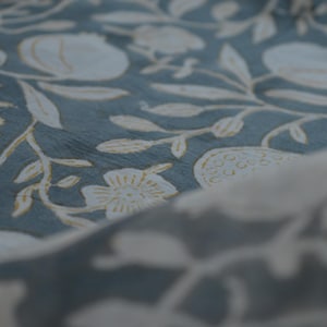 May include: Close-up of a fabric with a repeating floral pattern. The design features white flowers, leaves, and fruit shapes on a muted teal background. The fabric has a soft, textured appearance.
