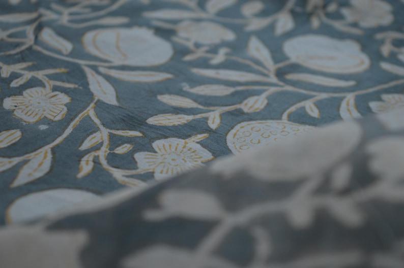 May include: Close-up of a fabric with a repeating floral pattern. The design features white flowers, leaves, and fruit shapes on a muted teal background. The fabric has a soft, textured appearance.