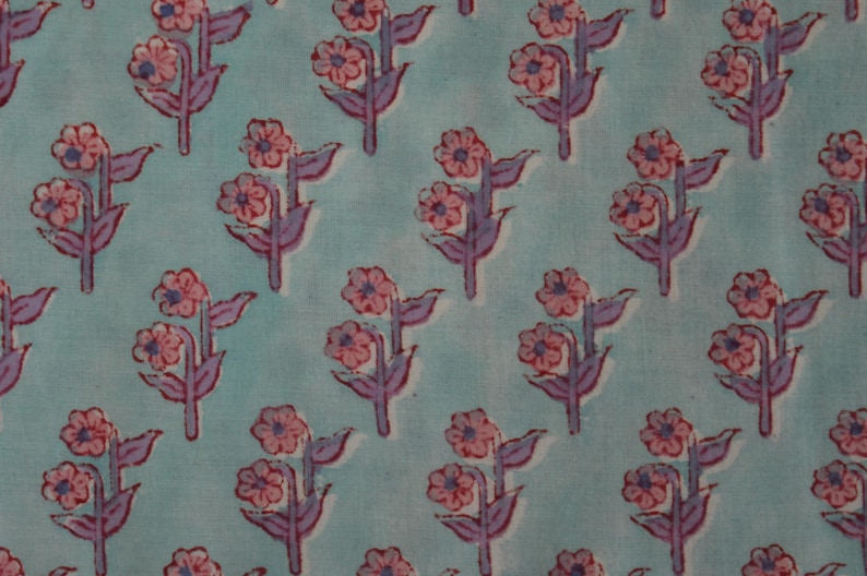 May include: A repeating pattern of stylized flowers on a light blue background. The flowers are pink with purple stems and leaves. The fabric has a soft, slightly faded appearance, suggesting a vintage or handmade quality.