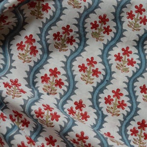May include: Close-up of a fabric with a repeating floral pattern. The design features red flowers with green stems and leaves, set against a white background. The pattern is framed by a wavy, light blue border.