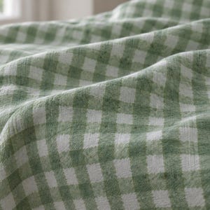 May include: A fabric textile featuring a sage green and white gingham check pattern. The material is draped with soft folds, displaying a woven texture with a consistent grid design throughout the surface of the cloth.