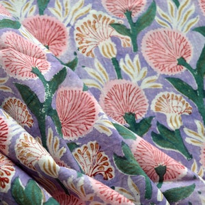 Puede incluir: Primer plano de una tela con un estampado floral. El diseño presenta flores rosas y blancas con tallos y hojas verdes sobre un fondo lavanda. La tela está doblada, mostrando el diseño repetitivo del patrón.