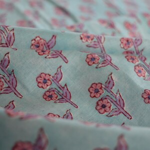 May include: Close-up of a light blue fabric with a repeating floral pattern. The flowers are pink with blue centers and purple stems and leaves. The fabric is slightly wrinkled, creating shadows and depth.