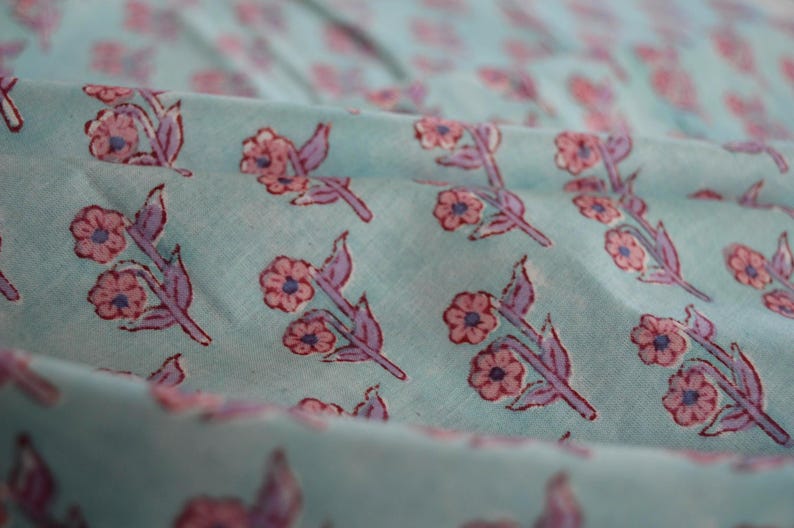 May include: Close-up of a light blue fabric with a repeating floral pattern. The flowers are pink with blue centers and purple stems and leaves. The fabric is slightly wrinkled, creating shadows and depth.
