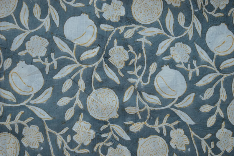 May include: A close-up of a fabric with a repeating pattern of white pomegranates, flowers, and leaves on a muted teal background. The design has a hand-drawn aesthetic, with visible texture and slight variations in the print.