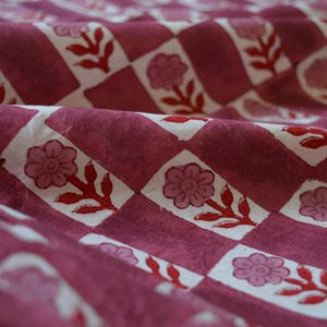 May include: Close-up of a fabric with a repeating pattern of flowers and leaves in red and white on a burgundy background. The fabric is folded, showing the texture and depth of the material.