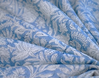 May include: Light blue fabric with a white floral pattern. The fabric is draped, showing the texture and design of the print. The floral design includes leaves and flowers.