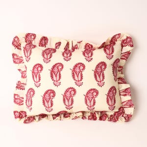 May include: Cream-coloured rectangular cushion featuring a repeating paisley pattern in red hues. The cushion has a ruffled edge with a matching paisley print. A classic decorative item for home interiors.