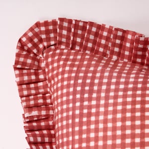 Op de afbeelding: Close-up van een kussen met een rood-wit ruitpatroon. De rand van het kussen is afgewerkt met een ruche. De geruite stof creëert een gezellige en uitnodigende sfeer.
