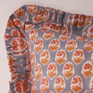 Puede incluir: Un cojín decorativo con un estampado floral en tonos naranja, blanco y gris. El cojín tiene un borde con volantes y un patrón repetido de flores estilizadas. La tela parece ser de algodón o lino.