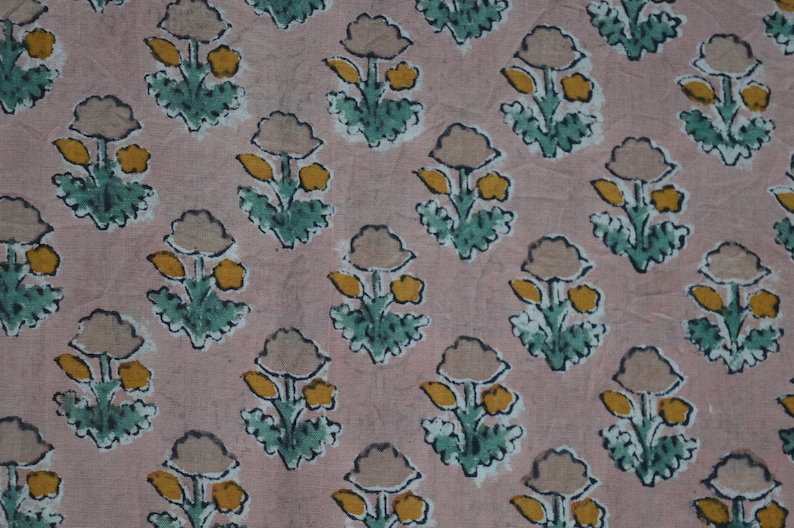 May include: A fabric with a repeating floral pattern. The design features stylized flowers in shades of beige, yellow, and green, set against a soft pink background. The flowers have a simple, block-print style, creating a vintage aesthetic.