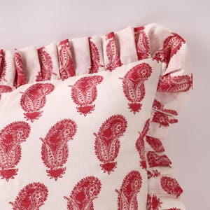 May include: A white pillow with a red paisley print and a ruffled edge. The pillow is made of cotton and is soft and comfortable.