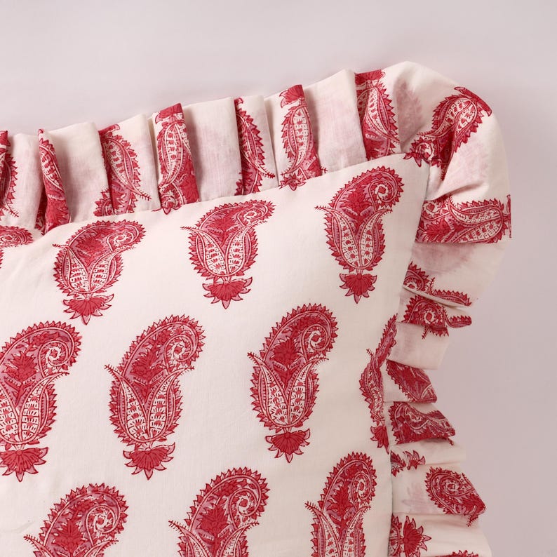 May include: A white pillow with a red paisley print and a ruffled edge. The pillow is made of cotton and is soft and comfortable.