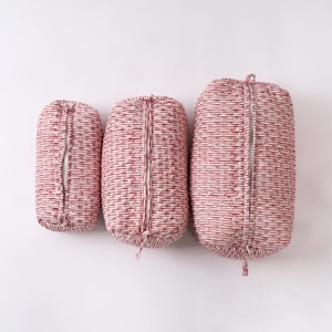 Pode incluir: Três bolsas com zíper de tecido vermelho e branco de tamanhos variados. As bolsas são cilíndricas com um zíper vertical no centro. O tecido tem uma aparência texturizada com um padrão repetido de linhas vermelhas e brancas.